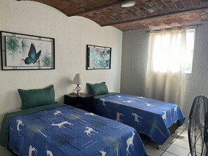 Una habitación con dos camas, una mesita de noche con lámpara, ventilador de techo y obras de arte en las paredes.