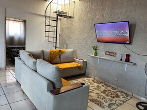 Una sala de estar con un sofá, un televisor montado en la pared, un estante con adornos y una alfombra estampada.