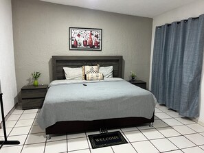 A neatly arranged bedroom with a bed, a bedside table with a plant, a framed artwork on the wall, and a welcome mat on the floor.