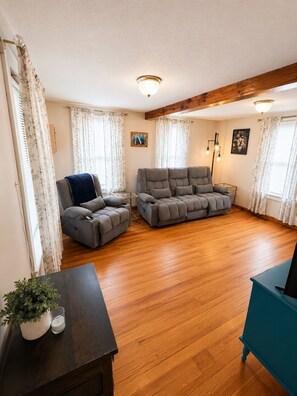 Ein geräumiges Wohnzimmer mit Holzboden, einem grauen Sofa, einem blauen Sessel und einem dunklen Holztisch mit Pflanze und Kerze.