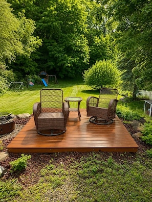 Eine Holzterrasse mit Korbstühlen und einem kleinen Tisch, umgeben von üppiger Bepflanzung und einem Spielplatz im Hintergrund.