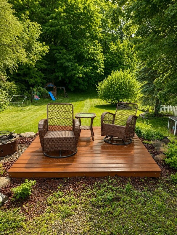 A wooden deck with wicker chairs and a small table, surrounded by lush greenery and a playground in the background.
