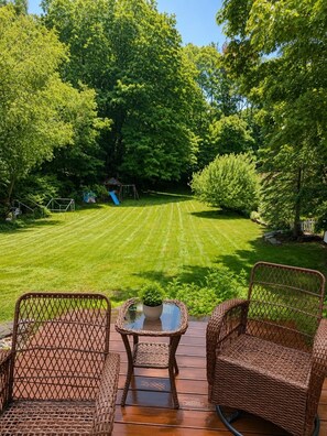 A deck with wicker furniture, a small table with a plant, and a view of a well-maintained lawn and trees.