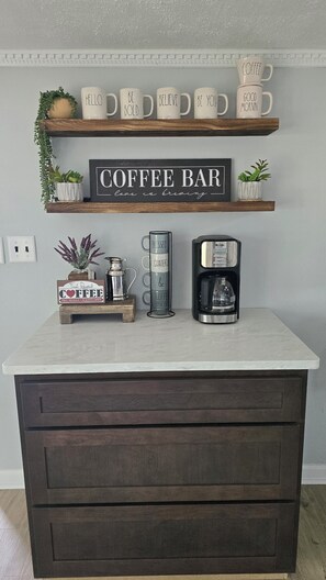 A kitchen counter with a coffee maker, a sign that says 'Coffee Bar,' and a shelf with coffee mugs.