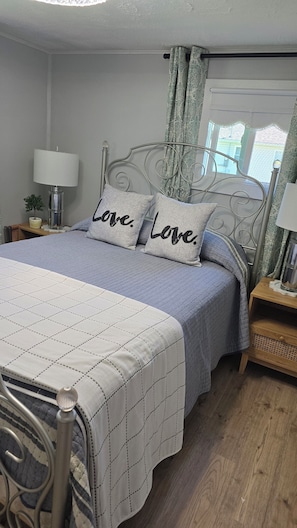 A bedroom with a metal bed frame, a white quilted bedspread, two pillows with 'Love.' text, a bedside lamp, a small plant, and a window with patterned curtains.