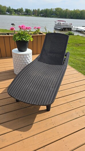A deck with a wicker lounge chair, a hexagonal white planter with pink flowers, and a view of a lake with a boat in the background.