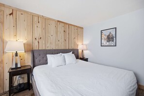 A hotel room with a wooden paneled wall, a bed with white bedding, two bedside lamps, and a framed picture of a snow train. - Eastern Inn Snowy Ridge Suite (North Conway)
