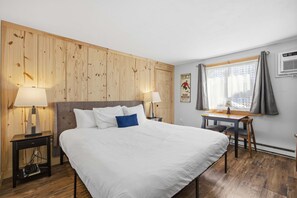 Ein Schlafzimmer mit einem Bett, zwei Nachttischen mit Lampen, einem Schreibtisch mit Stuhl, einem Fenster mit Vorhängen und einer Wand-Klimaanlage.