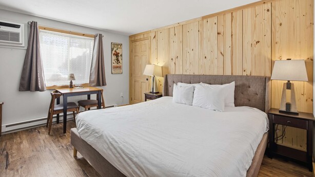 A bedroom with a bed, a desk, two lamps, a window with curtains, and a wooden wall panel. - Eastern Inn Mountain Ski Lodge (North Conway)