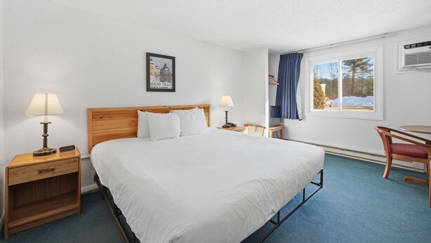 Une chambre d’hôtel avec un lit, deux tables de chevet avec des lampes, une petite table avec une chaise, une fenêtre donnant sur un paysage agréable et une affiche accrochée au mur.