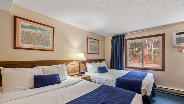 A hotel room with two beds, a nightstand with a lamp, framed artwork on the walls, a window with curtains, and an air conditioning unit. - Eastern Inn Hemlock Hideout (North Conway)