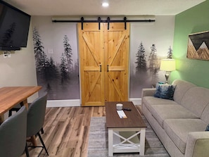A room with a wooden dining table, a sofa, a TV, and a sliding door with a forest mural. - Eastern Inn Evergreen Suite (North Conway)