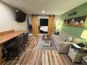 A room with a wooden dining table, a sofa, a TV mounted on the wall, and a bed with a view of the mountains. - Eastern Inn Evergreen Suite (North Conway)