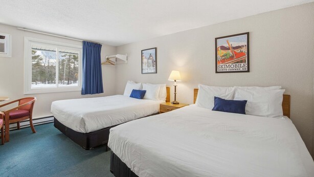 Une chambre d’hôtel avec deux lits, un bureau, une chaise, une fenêtre avec des rideaux, une lampe et une affiche au mur.