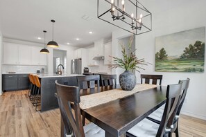 A modern kitchen with white cabinets, a central island, pendant lighting, and a dining area with a wooden table and chairs. - Birdsong Under the Stars | Hot Tub & Fire Pit! (Fredericksburg)