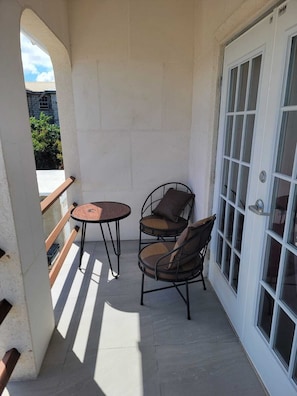 Ein kleiner Balkon mit Tisch und zwei Stühlen, eine weiße Tür mit Glasfeldern und Blick auf ein Gebäude im Hintergrund.