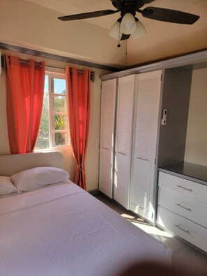 Ein Schlafzimmer mit einem Bett, einem Deckenventilator, einem Fenster mit Vorhängen und einem Kleiderschrank.