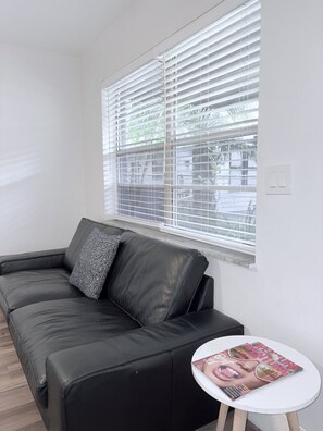 A black leather sofa with patterned cushions, a small white table with a magazine, and a window with horizontal blinds. - Sailboat Bend 1BR | 2 Queen Beds | Free Parking | Near Las Olas (Fort Lauderdale)