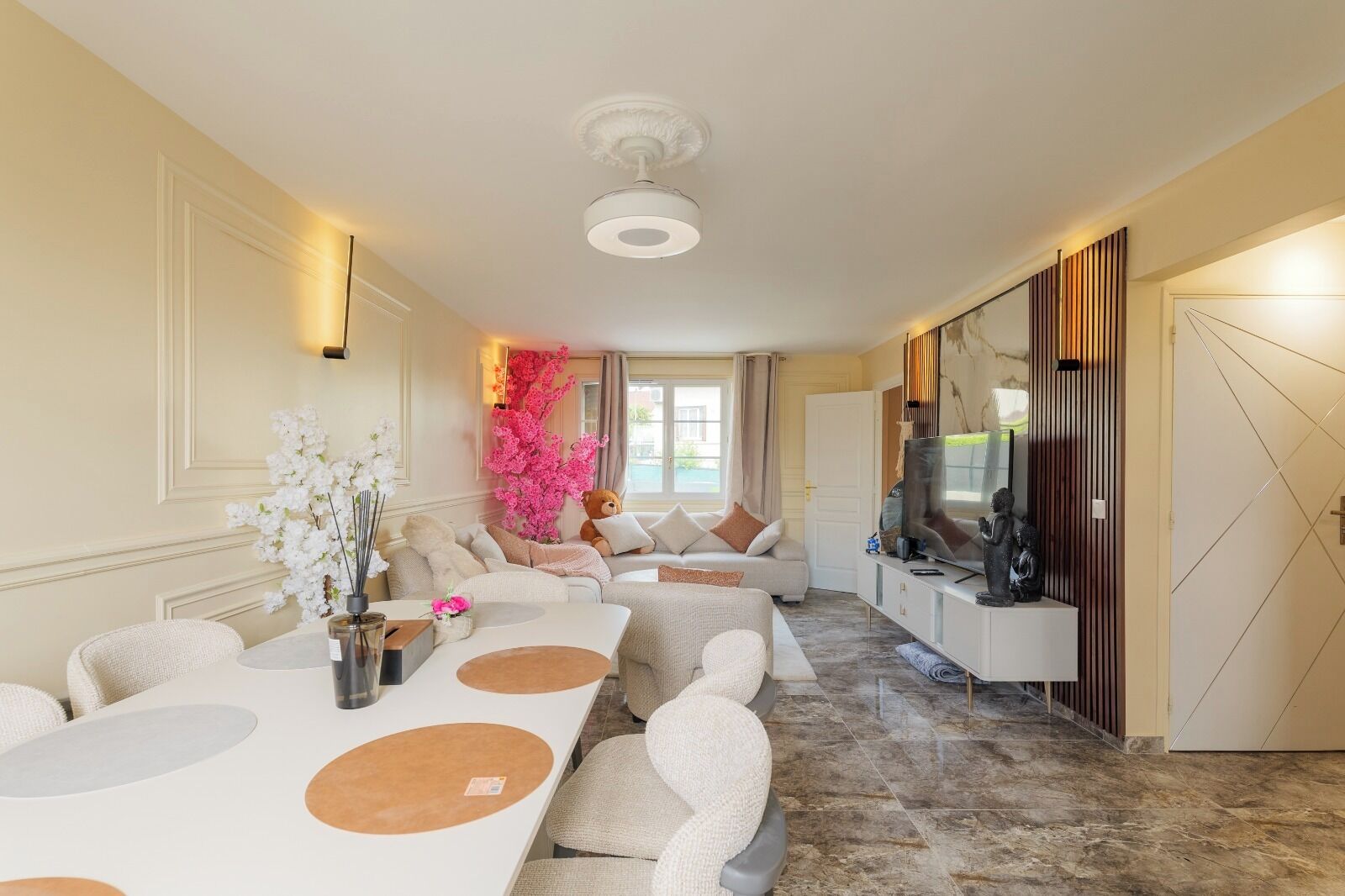 Een ruime kamer met een lange eettafel, banken, een televisie en een decoratieve, roze plant.