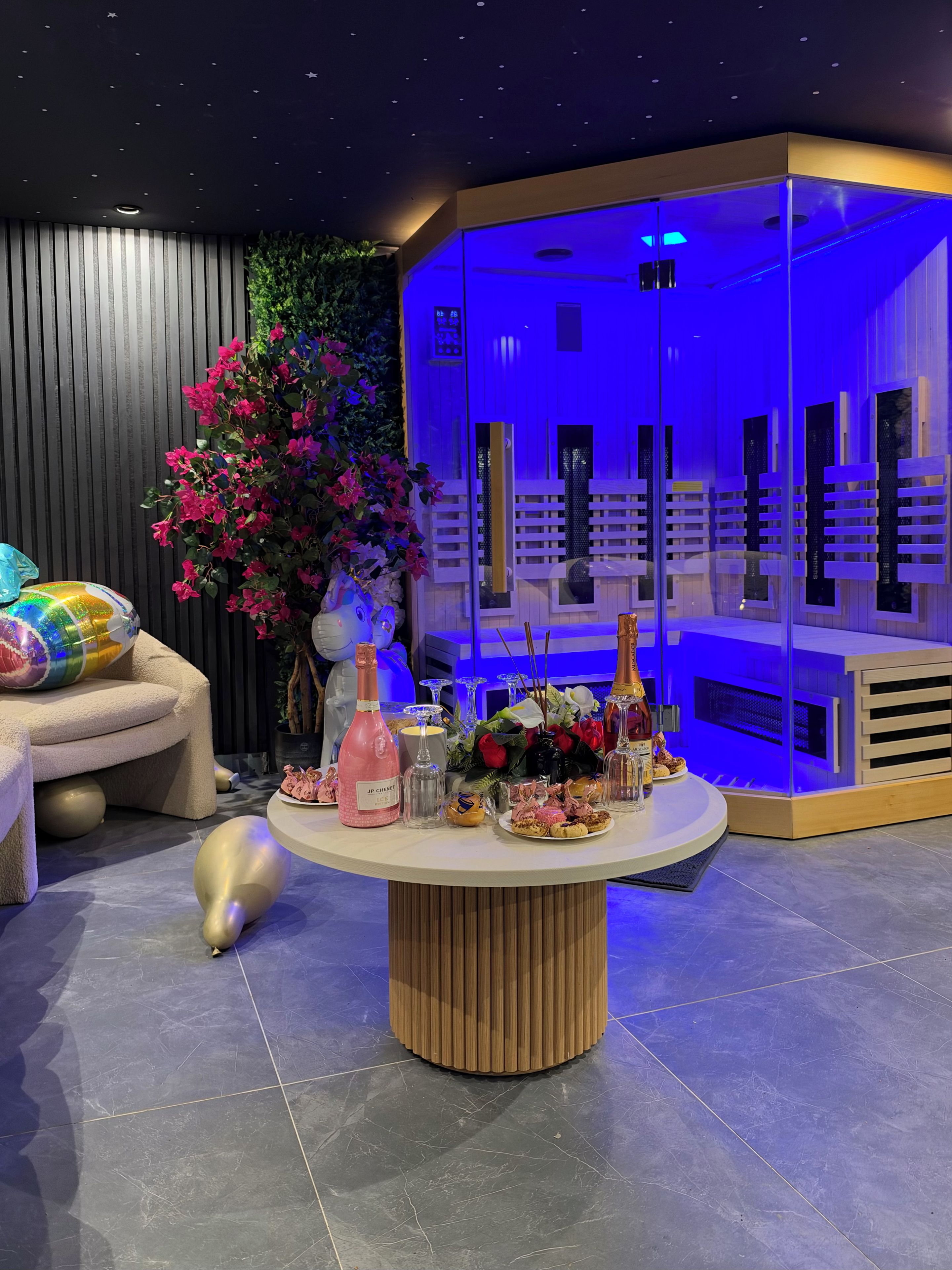 A modern interior with a round table displaying various items, a seating area with a sofa and decorative pillows, and a backdrop with a vertical garden and a blue-lit structure.