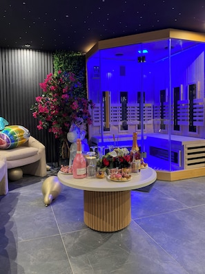 Un interno moderno con un tavolo rotondo su cui sono esposti diversi oggetti, una zona giorno con divano e cuscini decorativi, e uno sfondo caratterizzato da un giardino verticale e una struttura illuminata di blu.