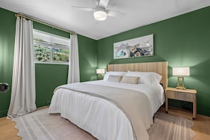 Ein Schlafzimmer mit Bett, zwei Nachttischen, einem Deckenventilator, einem Fenster mit Vorhängen und einem Wandbild.