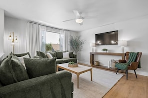 Ein Wohnzimmer mit einer Couch, einem Couchtisch, einem Fernseher, einem Deckenventilator und einem Fenster mit Vorhängen.