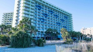 Un hotel grande y de varias plantas con diseño moderno, que cuenta con balcones en cada piso y está ubicado en primera línea de playa, con palmeras.