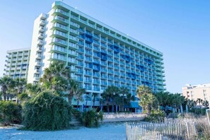 A large, multi-story hotel with a modern design, featuring balconies on each floor and a beachside location with palm trees. - Oceanfront Unit with Private Balcony and Pool Access (Myrtle Beach)