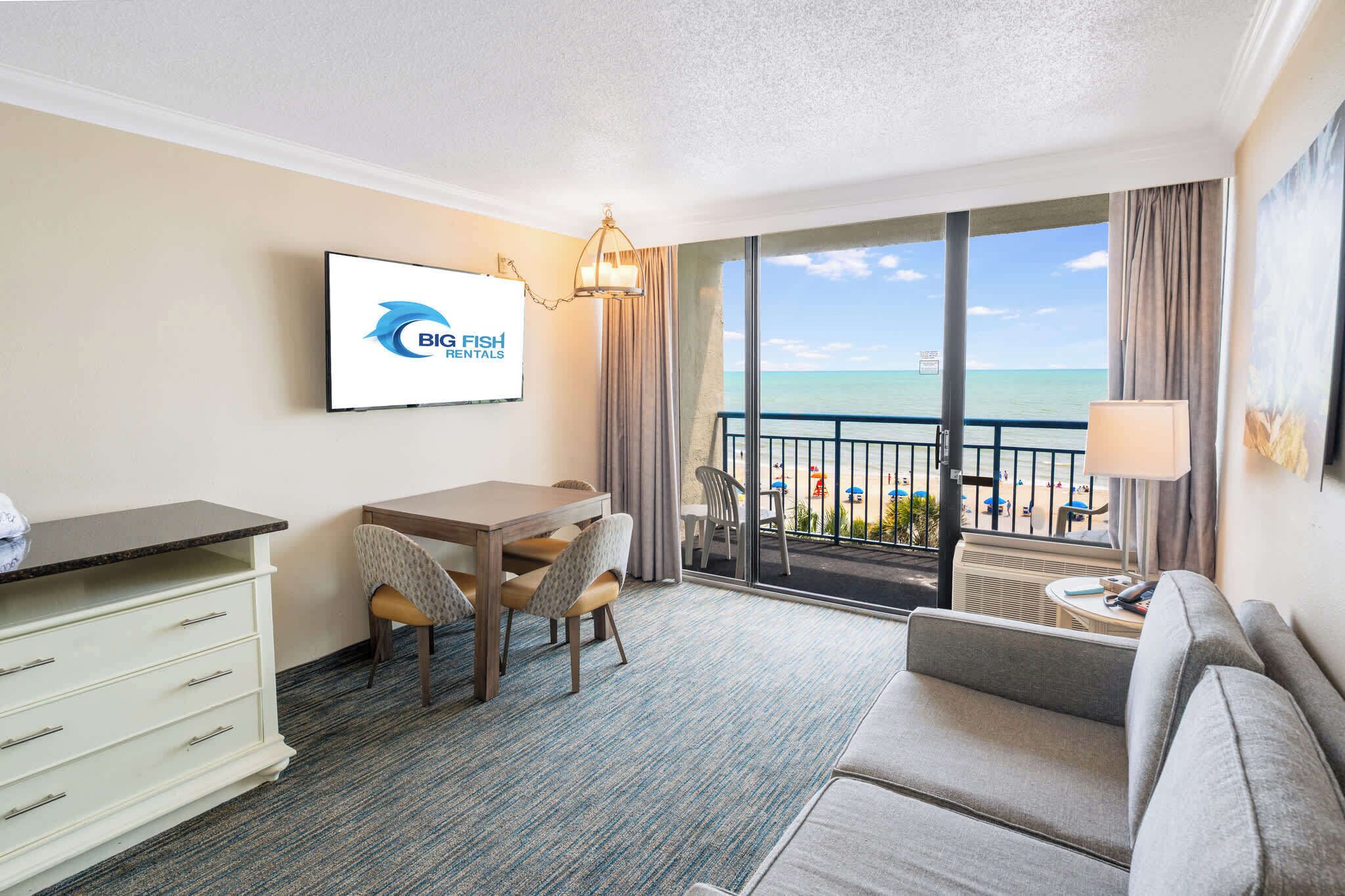 Habitación de hotel con balcón, zona de comedor, sofá y televisor mostrando 'BIG FISH RENTALS'.