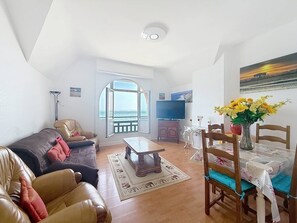 Ein Wohnzimmer mit einer Couch, einem Couchtisch, einem Esstisch mit Stühlen, einem Fernseher und Meerblick durch ein großes Fenster.