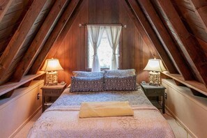 A cozy attic bedroom with a sloped ceiling, a bed with patterned bedding, two bedside lamps, and a window with curtains.