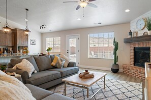 A living room with a sofa, coffee table, and fireplace. There is a kitchen area with cabinets and a countertop. - Stunning 5BR Oasis • Hot Tub • Casita • Hurricane (Hurricane)
