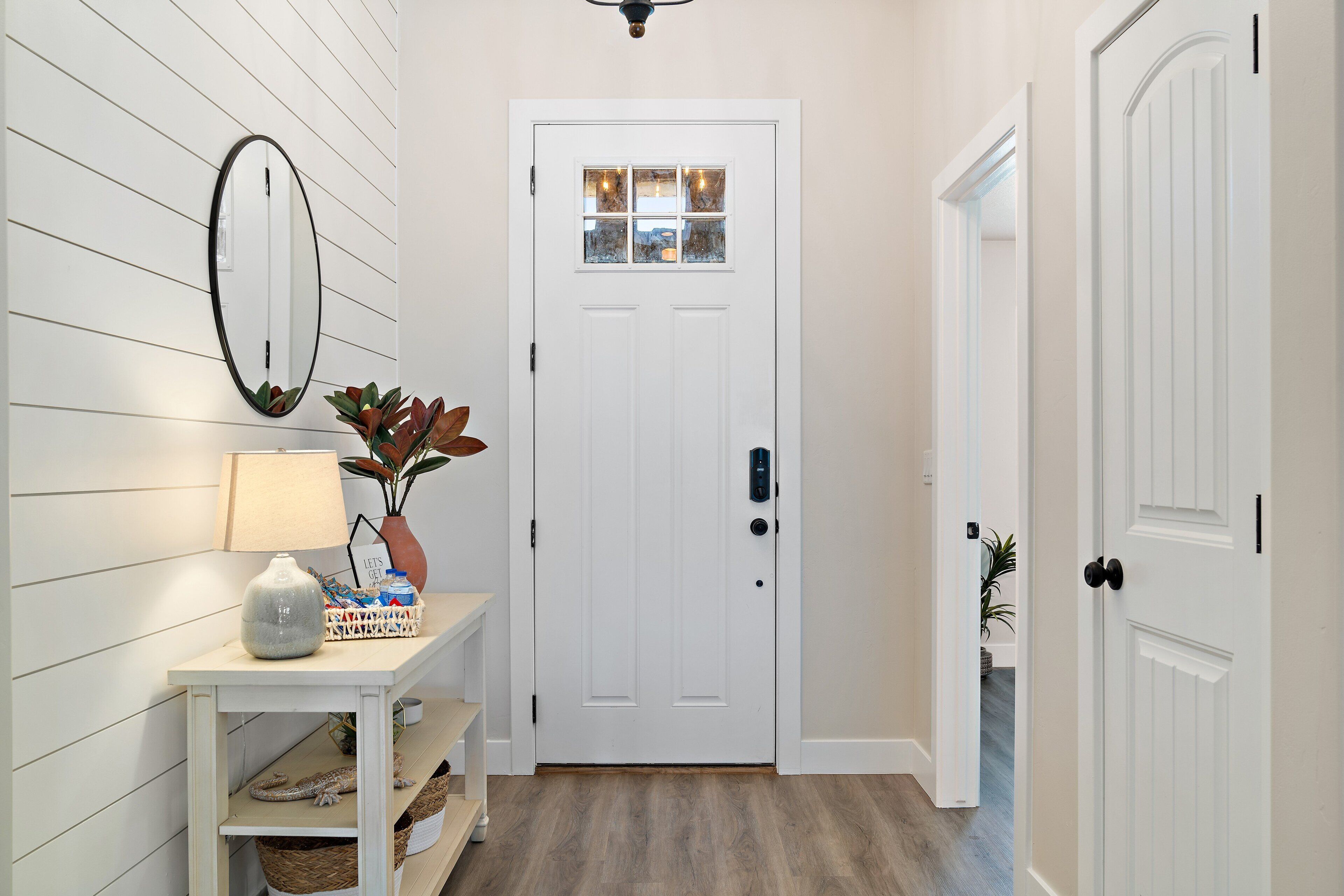A hallway with a white door, a mirror, a lamp, and a small table with a vase and a basket.