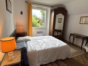 Ein Schlafzimmer mit einem Bett, einem Nachttisch mit Lampe, einem Fenster mit Vorhängen, einem Spiegel und einem kleinen Tisch.
