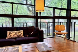 Ein Zimmer mit einem dunkelgrauen Sofa, einem Holzstuhl, einem Tisch mit Büchern und großen Fenstern mit Blick auf Bäume und einen Zaun.