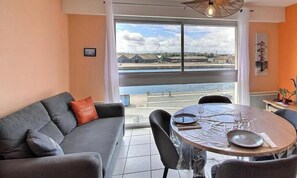 Ein modernes Wohnzimmer mit grauem Sofa, einem runden Esstisch und Blick auf ein Gewässer durch große Fenster.