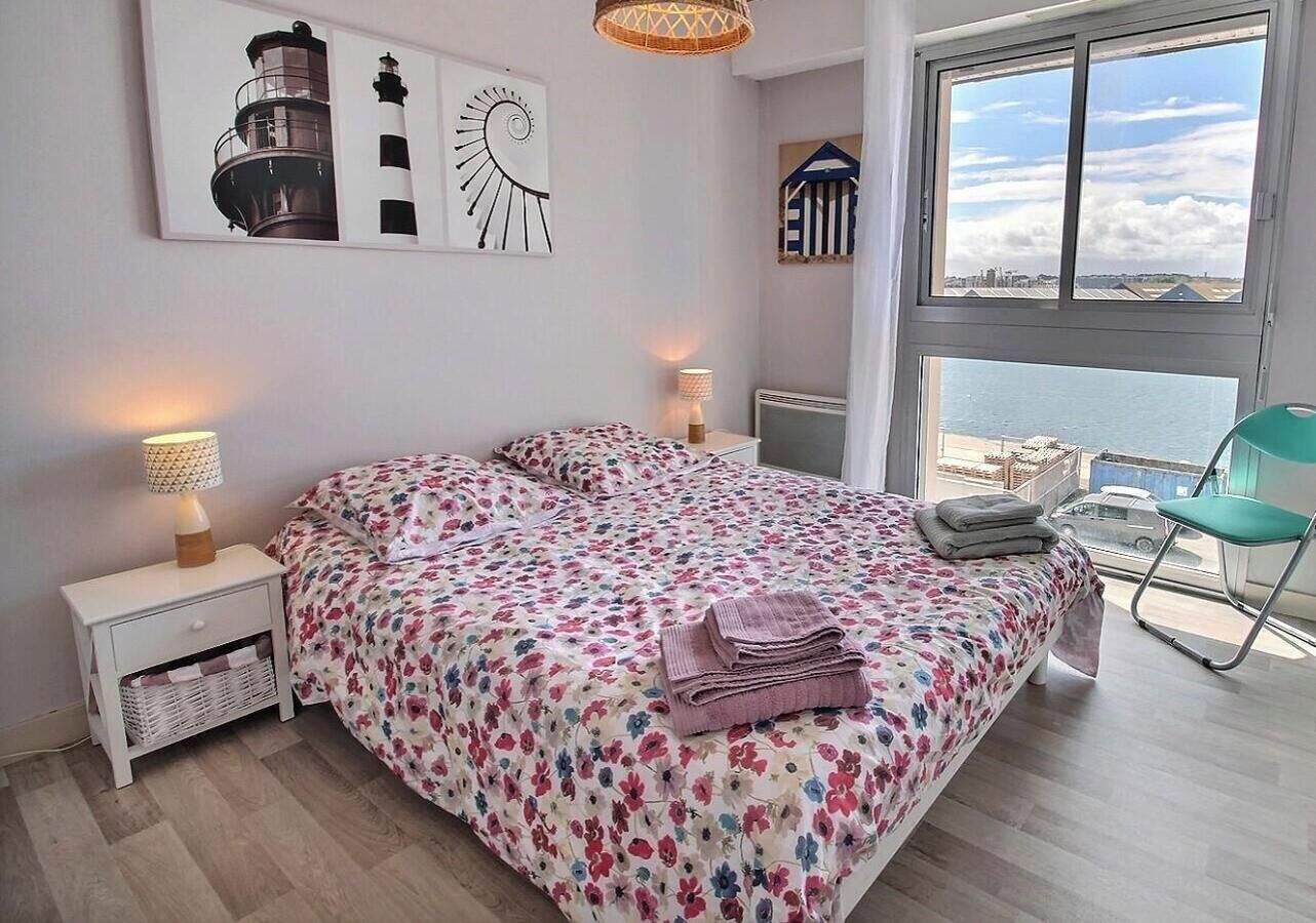 Un dormitorio con cama, mesita de noche, silla, ventana con vistas y decoración en la pared.