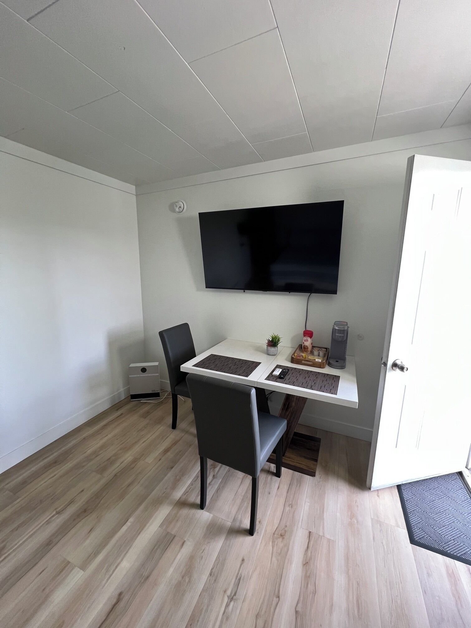 A room with a white desk, a black chair, a flat-screen TV mounted on the wall, a small potted plant, and a white door.