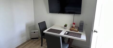 A room with a white desk, a black chair, a flat-screen TV mounted on the wall, a small potted plant, and a white door.