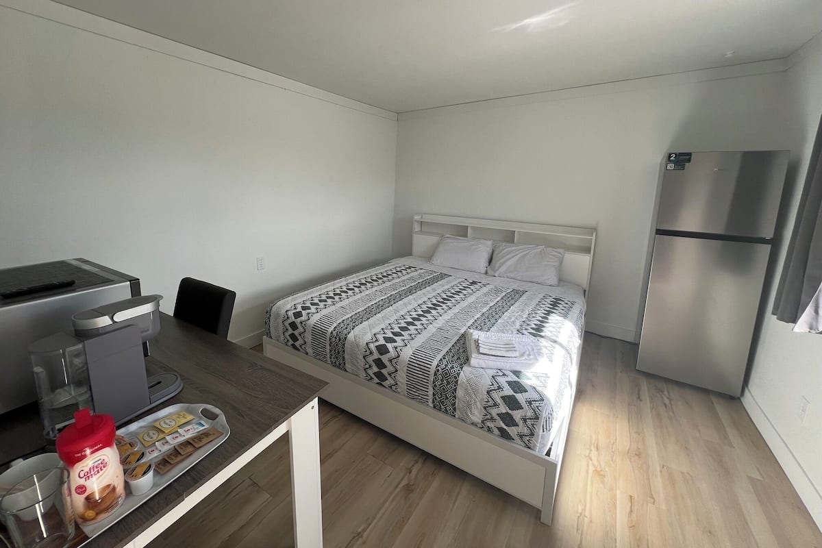 A bedroom with a bed, a desk with a computer, a coffee maker, and a refrigerator.
