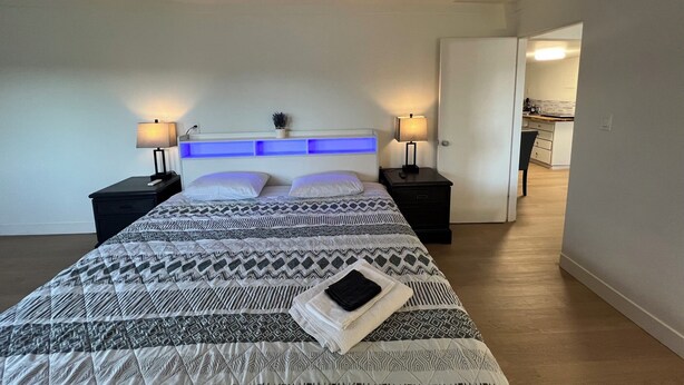 Un dormitorio moderno con una cama grande, mesitas de noche, un cabecero con iluminación azul y vista a la cocina.