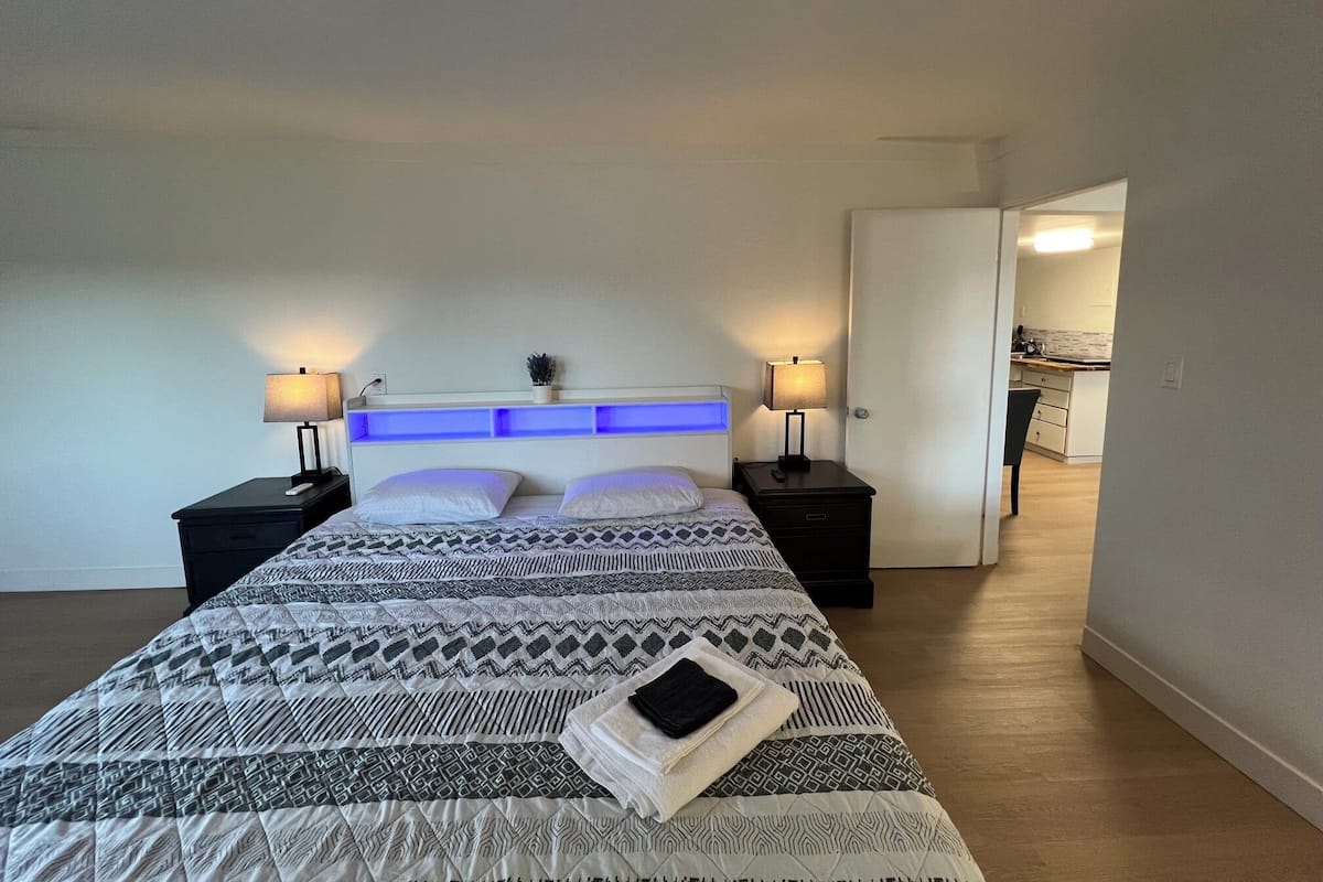 A modern bedroom with a large bed, bedside tables, a headboard with blue lighting, and a view into the kitchen.