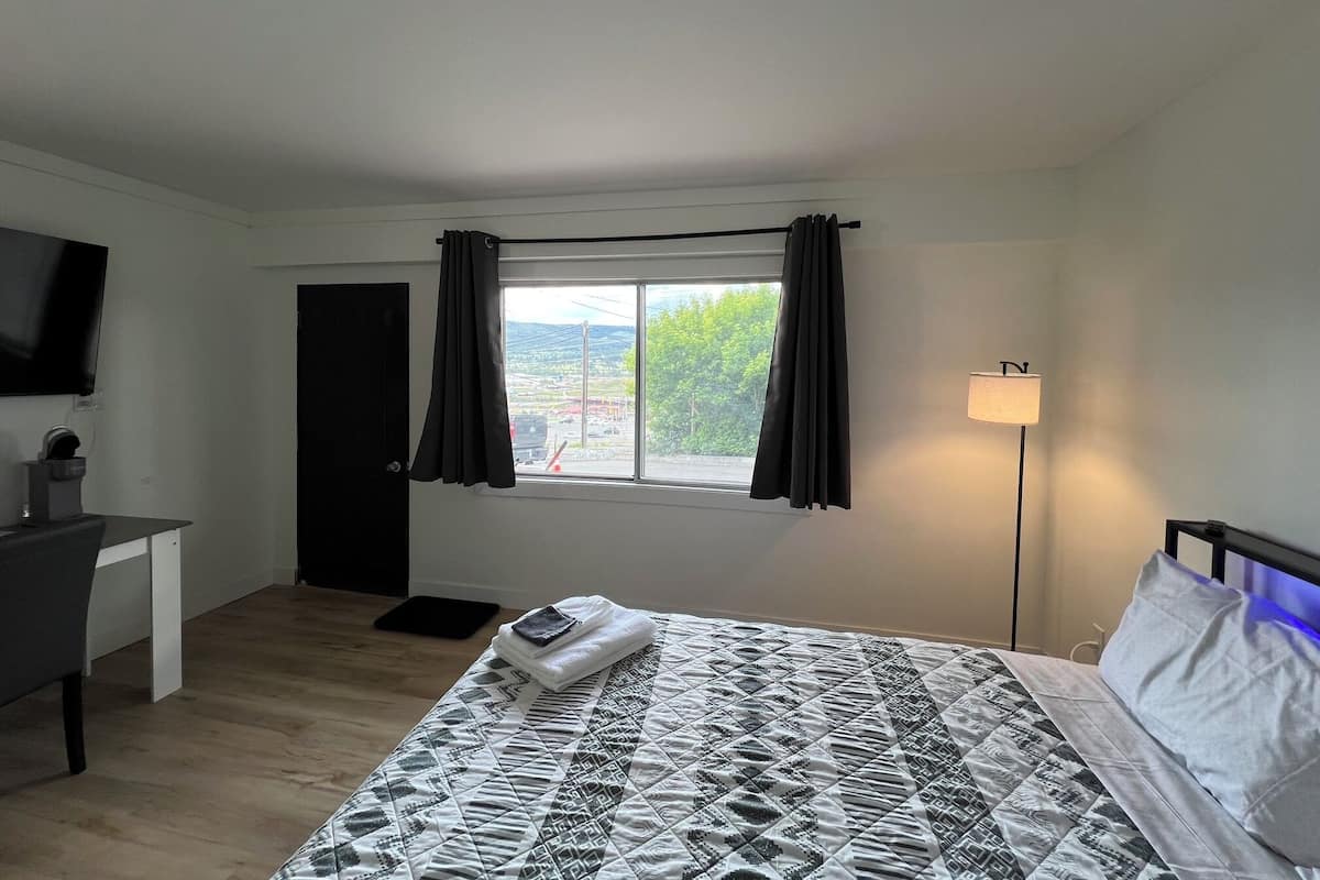 A bedroom with a bed, a desk, a TV, a window with curtains, and a lamp.