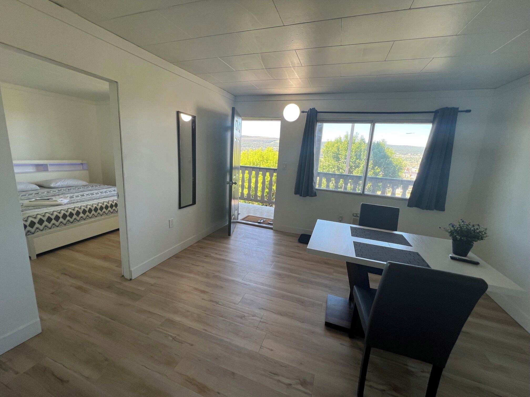 Una habitación con cama, escritorio con silla, planta en maceta y ventana con vista a follaje.