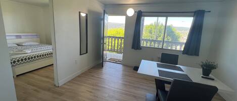 Una habitación con cama, escritorio con silla, planta en maceta y ventana con vista a follaje.