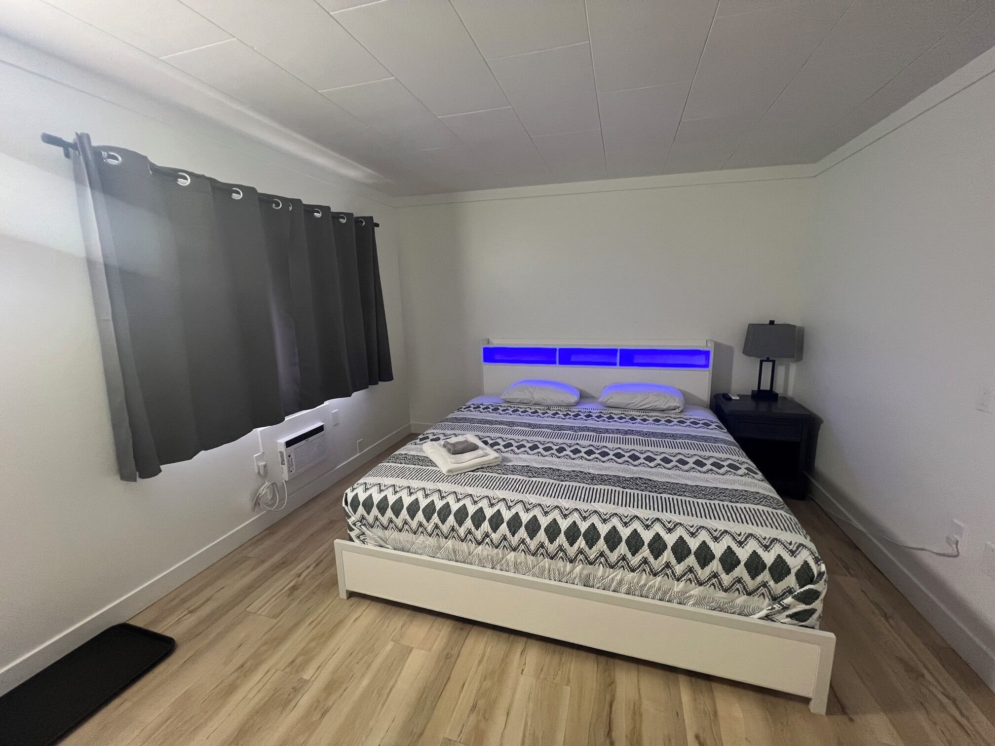 A bedroom with a bed, a bedside table with a lamp, a window with curtains, and a wall-mounted heater.