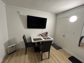 Una habitación pequeña con una televisión blanca montada en la pared, un escritorio con dos sillas, un calentador pequeño y una puerta con luz.