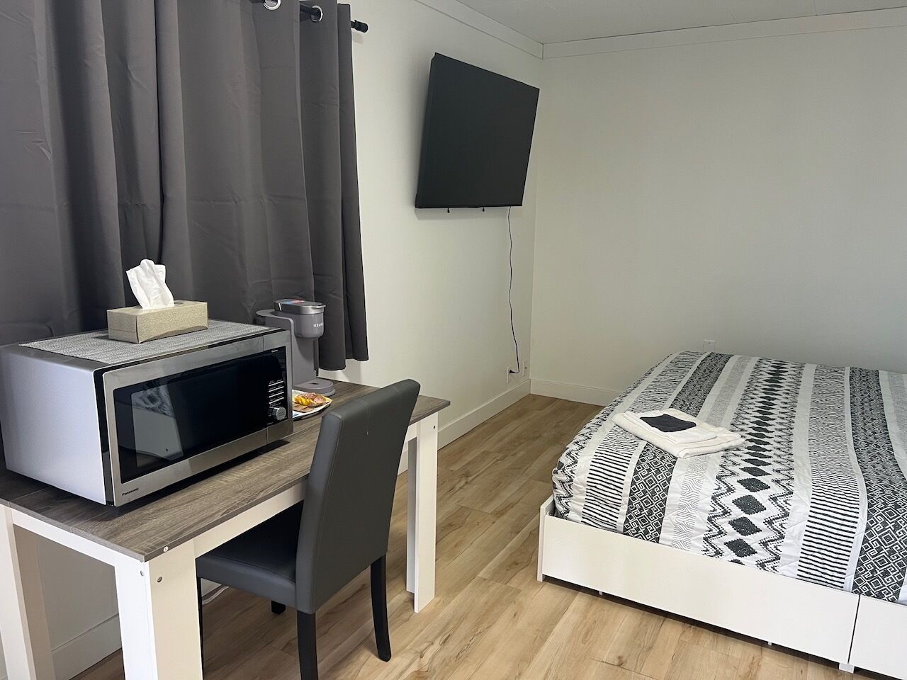 Ein kleines Zimmer mit einem Bett, einem Schreibtisch mit einer Mikrowelle, einem Stuhl, einem an der Wand befestigten Fernseher und einem Vorhang.
