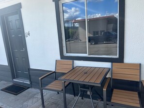 A small outdoor seating area with a table and two chairs, a black door with the number 114, and a window overlooking a street.